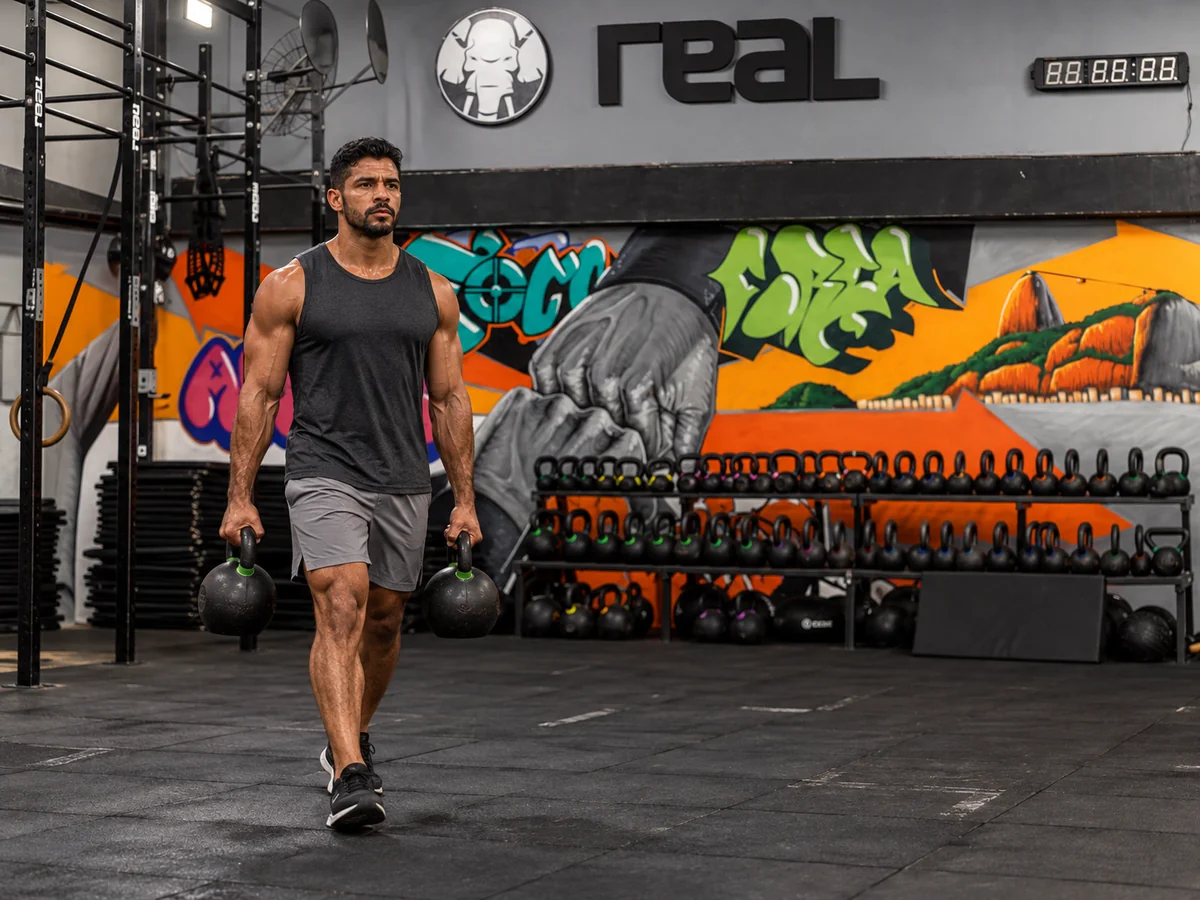 Homem de porte atlético caminhando com kettlebells na Real Cross — imagem ilustrativa do Híbrido